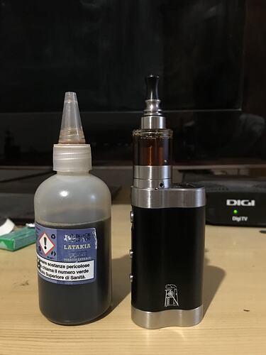 A black-silver electric cigarette and a bottle containing a dark liquid, labeled "Latakia Tobacco Extract" are visible on a table, with a TV and a Digi TV box in the background. (Subtitled by AI)