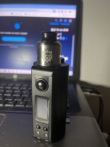 A black vape device stands in front of a laptop keyboard, the laptop screen lights up from behind. (Captioned with AI)