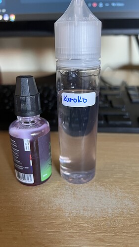On the table, in front of a keyboard, stand a small bottle with a black cap containing purple liquid and a larger plastic bottle with a label reading "Kuroko" containing clear liquid. (Labeled by AI)
