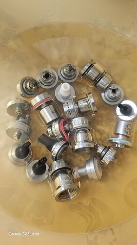 The image shows various metal vape tank parts in a clear container. (AI-captioned)