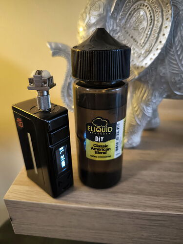 A black box mod with an atomizer on top, and a dark e-liquid bottle stand on a wooden surface, with a silver elephant statue in the background. (AI-captioned)