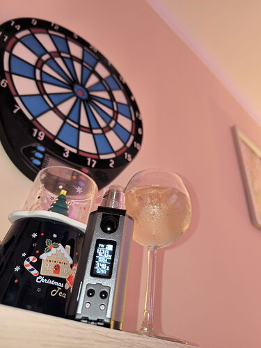 A rĂłzsaszĂn falon egy dartstĂĄbla alatt egy polcon karĂĄcsonyi tĂ©mĂĄjĂș bögre, egy vape eszköz Ă©s egy italos pohĂĄr lĂĄthatĂł. (AI-feliratozott)