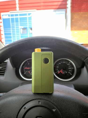 A green electric cigarette is placed on a car's steering wheel, with the car's dashboard and a building in the background. (Captioned by AI)