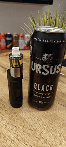 A black vape and an Ursus Black beer can stand on a wooden table, with plants and more cans visible in the background. (Captioned by AI)