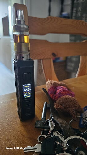 An electronic cigarette sits on a table with a wooden chair in the background, keys and a knitted item are visible next to it. (AI-captioned)