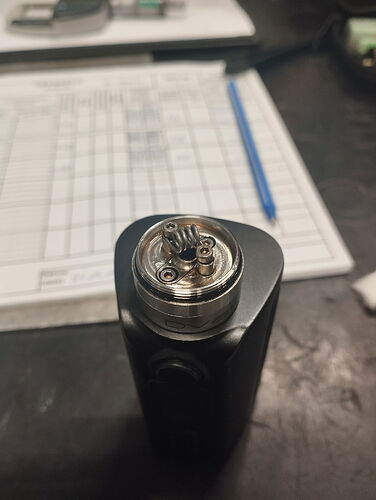 The image shows a close-up of an electric Vape device with a constructed stainless steel coil, a writing instrument and a tabular document in the background. (Captioned by AI)