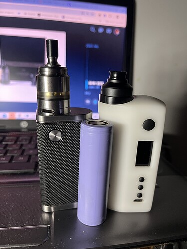 A képen egy fekete és egy fehér vape készülék, valamint egy lila elem látható, háttérben egy laptop képernyőjével. (Feliratozva AI-val)