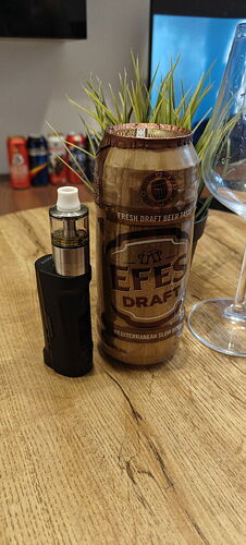 A black vape device and an Efes Draft beer can are seen on a wooden table, accompanied by a plant, a wine glass, and additional drink cans in the background. (Captioned by AI)