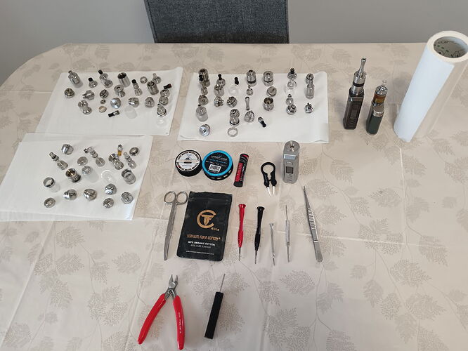 Various vape tank parts, tools, and two electronic cigarette mods are arranged on the table. (Labeled by AI)