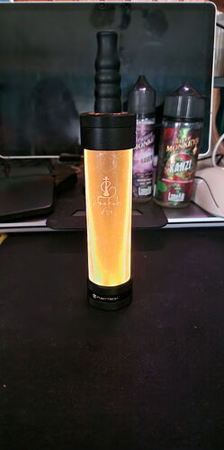 In the picture, a glowing, orange lamp is visible, which has a black base and top, and in the background, there are bottles filled with various liquids. (Transcribed by AI)