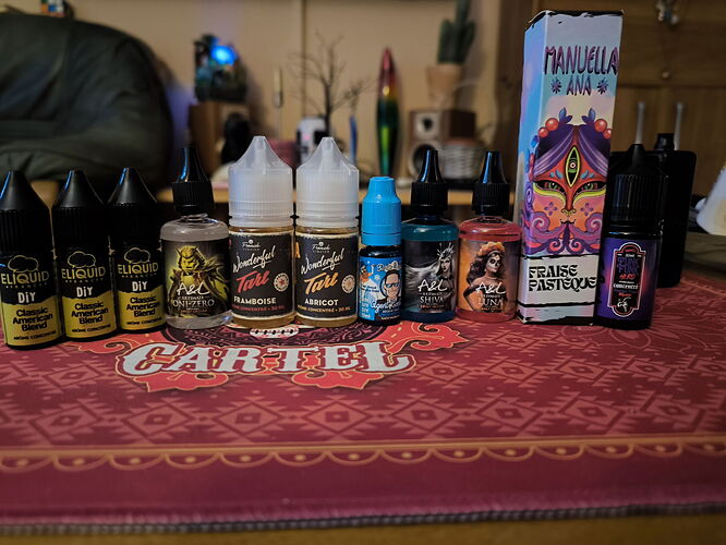 Various e-liquid bottles and boxes are visible on the table on top of a red coaster with the inscription CARTEL, with furniture in the background. (Caption by AI)