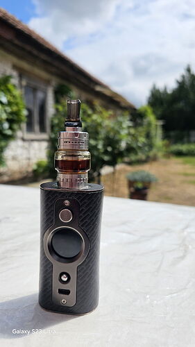 A black, textured electronic vape device is seen on a white table, with an old stone house and green vegetation in the background. (Caption by AI)