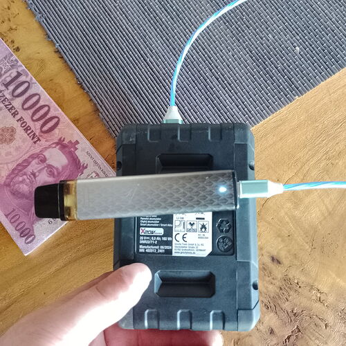 Egy személy egy kézben tartott eszközön keresztül USB-kábellel tölt egy modern elektromos cigarettát egy fekete töltőállomáson, mellette pedig egy 10 000 forintos bankjegy látható egy faasztalon. (Feliratozva AI-val)