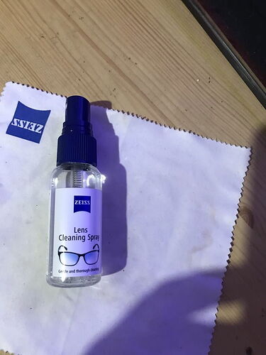 A ZEISS branded Lens Cleaning Spray bottle and a ZEISS-labeled microfiber cleaning cloth are visible on the table. (AI-generated caption)