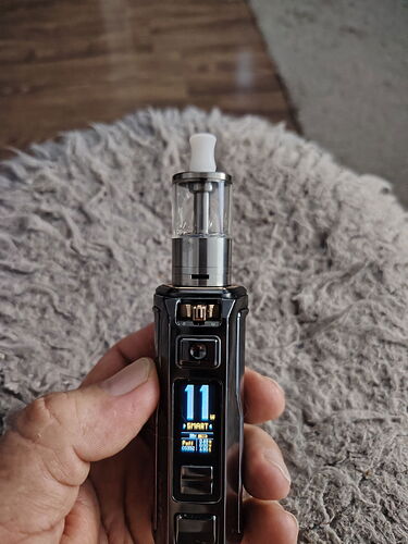 Egy kéz egy fekete elektromos dohányzóeszközt (vape-t) tart, amelyen digitális kijelző látható, a háttérben egy szőnyeg és fa padló található. (Feliratozva AI-val)