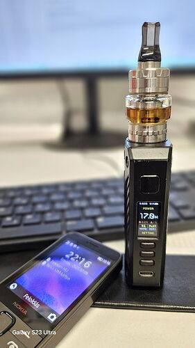 An electronic cigarette sits on a closed laptop on a table, with a mobile phone next to it, and a blurry computer screen and keyboard in the background. (Caption by AI)