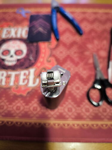 Az image egy felülről készült közelikép egy épített elektronikus cigaretta porlasztóról, amely egy piros mintás felületen helyezkedik el, háttérben ollóval és fogóval. (Feliratozva AI-val)