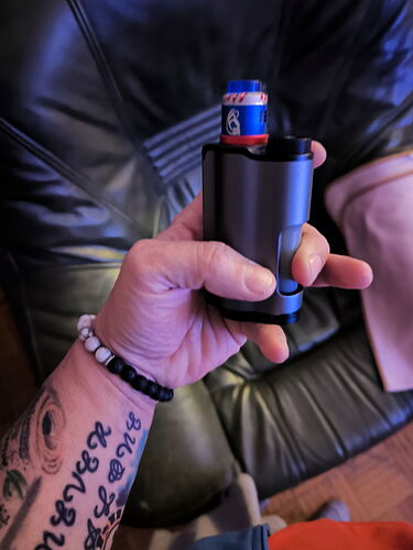 Une main tient un appareil de couleur noire et bleue, avec un canapé en cuir noir visible en arrière-plan. (Légendé par IA)