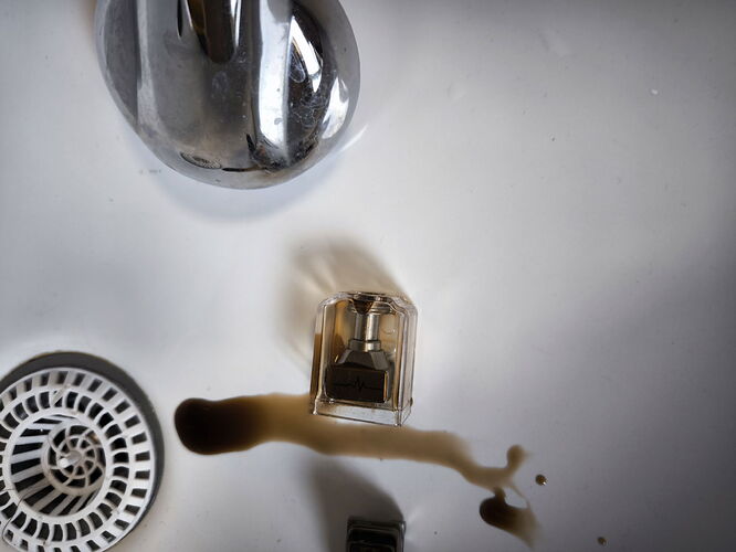 Une petite bouteille, dont un liquide sombre s'est renversé, repose dans un lavabo blanc près du drain et du robinet. (Légendé par IA)