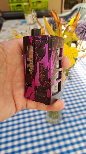 A hand holds a black and purple patterned electronic cigarette, with a blue-white checkered tablecloth and flowers in the background. (Labeled by AI)