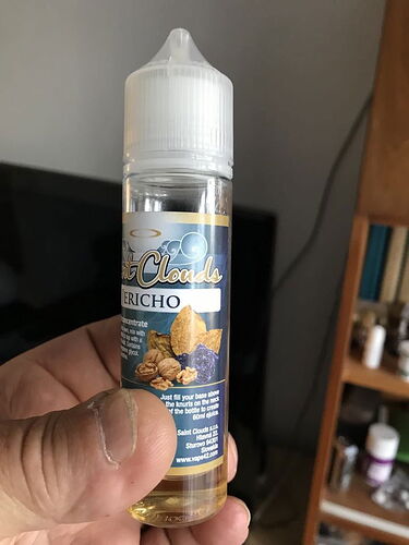 Une bouteille de concentré d'e-liquide, intitulée "Saint Clouds JERICHO", est tenue dans une main. Elle a une étiquette bleue et dorée ornée de noix et de feuilles. (Caption by AI)