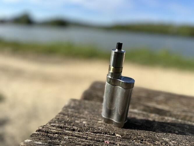 Un appareil de vape en métal posé sur le bord d'une planche en bois, sur fond de plage de sable et d'eau. (Légendé par IA)