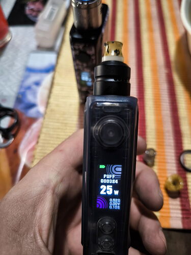 Egy kézben tartott, digitális kijelzős elektromos cigaretta eszköz egy asztal fölött, amelyen különböző tárgyak vannak elhelyezve. (Feliratozva AI-val)