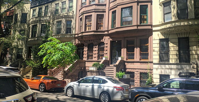 A sunny urban street scene where yellowish-brown old buildings and cars of various colors are parked along the sidewalk. (Captioned by AI)