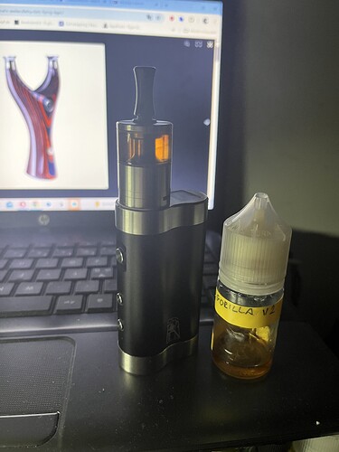 Une cigarette électronique noire et une petite bouteille contenant du liquide sur la table, avec une image de lance-pierre coloré visible à l'écran d'un ordinateur portable en arrière-plan. (Sous-titré par IA)