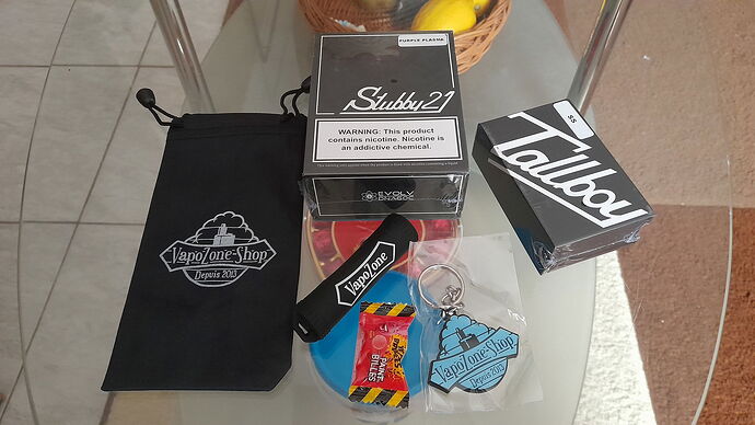 The image displays various vaping products and accessories on a glass table, including boxes labeled 'Stubby 21' and 'Tallboy', as well as a pouch and keychain with the 'VapoOne Shop' logo. (Labeled by AI)