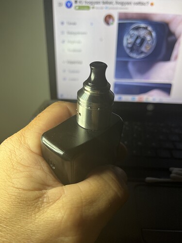 Egy kéz fekete vape eszközt tart, miközben egy laptop képernyője látható a háttérben. (Feliratozva AI-val)