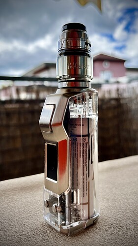 Egy elegáns, átlátszó és fém elektronikus cigaretta áll egy felületen, elmosódott épületek és felhős égbolt előtt. (Feliratozva AI-val)