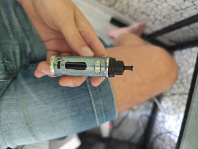 A hand holding a metal and plastic vape device, with a person wearing shorts in the background. (Caption by AI)