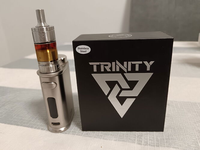 L'image montre une cigarette électronique argentée et une boîte noire avec l'inscription "TRINITY" et un logo géométrique. (Sous-titré par IA)