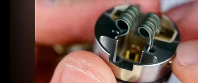 A képen egy kéz szorosan tart egy fém alkatrészt, amelyen két csavart alakú tekercs látható. (Feliratozva AI-val)