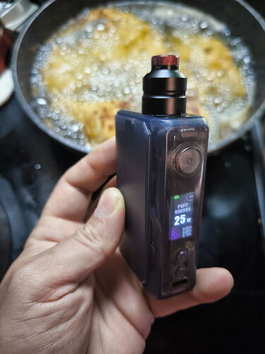 A person holds a black electric cigarette, while in the background, food is frying in hot oil in a pot. (Subtitled by AI)