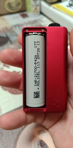 The image shows a hand holding a red metal housing battery holder, which contains a white Holicell INR-21700-P42A battery. (AI-generated caption)