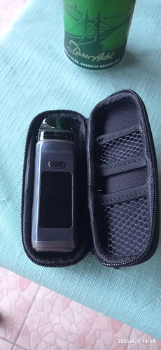 L'image montre un appareil électronique portable dans un étui noir ouvert, avec une boîte verte derrière lui sur une table. (Légendé par IA)