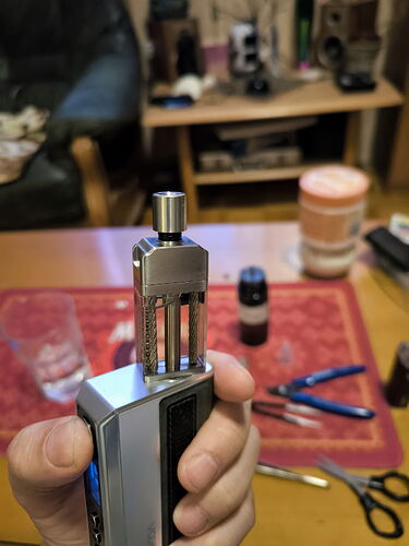 Une main tenant une cigarette électronique (vape), alors qu'à l'arrière-plan divers appareils et une table sont visibles. (Légendé par IA)