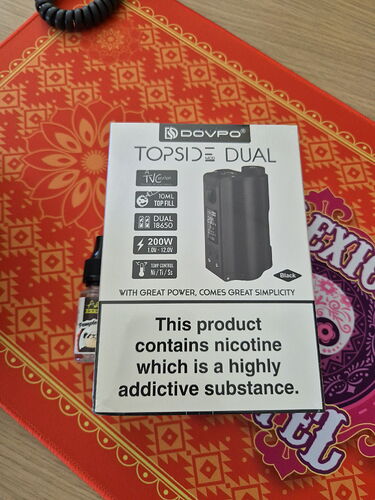 Dans l'image, la boîte d'une cigarette électronique Dovpo Topside Dual est visible, avec un avertissement de nicotine dessus, sur un dessous de plat rouge à motifs. (Légendé par IA)