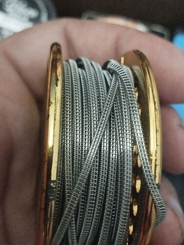 Une main tient une bobine dorée sur laquelle sont enroulés plusieurs fils métalliques filetés. (Étiqueté par l'IA)