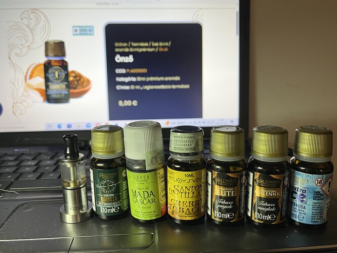 A képen hét különböző e-cigaretta folyadék üveg és egy porlasztó található egy laptop billentyűzetén, melynek képernyőjén egy illóolaj árusító weboldal látható. (Feliratozva AI-val)