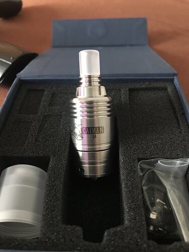 Un appareil de vapotage cylindrique de couleur argent, avec l'inscription Caiman V4, est montré dans une boîte à doublure en mousse bleue, avec d'autres accessoires à côté. (Légendé par IA)