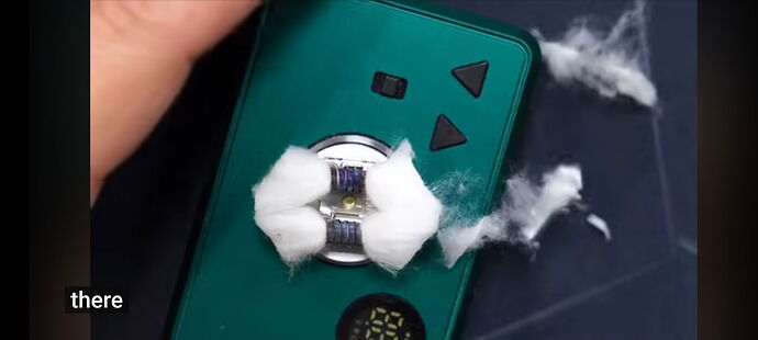 A hand holds a green vape device, with strands of cotton hanging out around the coils. (Captioned by AI)
