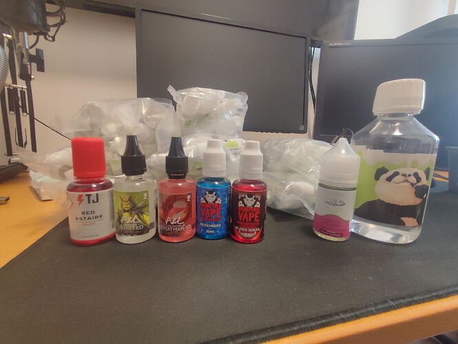 Divers flacons de e-liquides et une plus grande bouteille en plastique avec une image de panda sont visibles sur une table, avec des moniteurs et d'autres objets en arrière-plan. (Intitulé par IA)