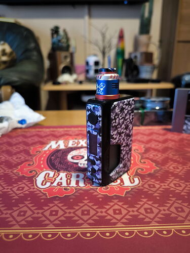 Az asztalon egy fekete-fehér mintás e-cigaretta készülék és a mögötte lévő tér elmosódott tárgyakkal látható. (Feliratozva AI-val)