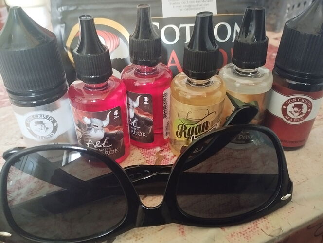 Sur la table, six bouteilles d'e-liquide de couleurs et d'étiquettes différentes, ainsi qu'une paire de lunettes de soleil noires sont visibles. (Légendé par l'IA)