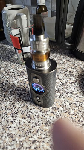 Une cigarette électronique argentée et foncée est visible sur un comptoir de cuisine tacheté, avec une tasse et d'autres objets en arrière-plan. (Légendé par l'IA)