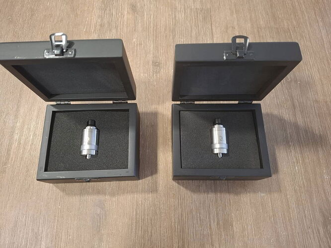 Two open black boxes are visible on a wooden surface, each containing a metal device embedded in black sponge. (Labeled by AI)