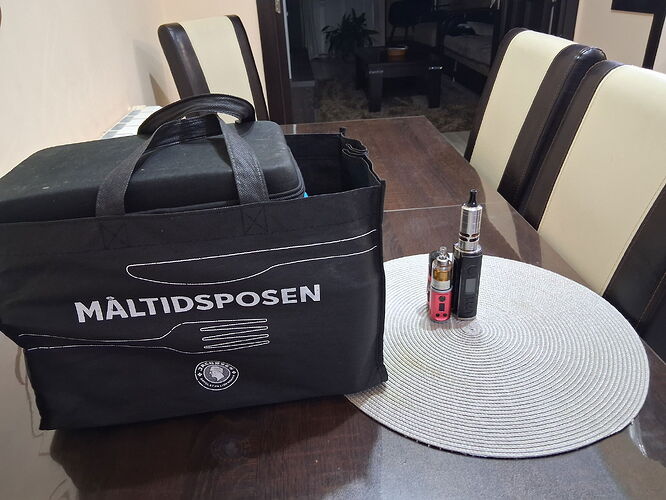 Sur la table se trouvent un sac noir marqué "MÁLTIDSPOSEN" et deux appareils de vapotage. (Légendé par IA)